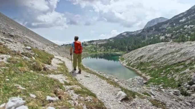 男性徒步旅行者在阳光下的山湖上方的砾石小径上行走