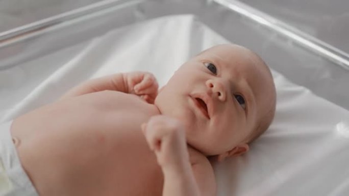 可爱的高加索新生婴儿躺在产科医院的摇篮里。一个顽皮而充满活力的孩子的特写脸肖像。医疗保健、怀孕和孕产