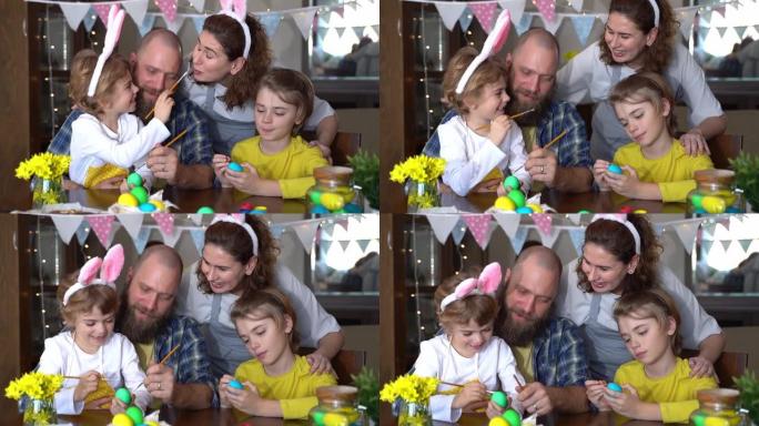 复活节家庭传统。父母和两个白种人快乐的孩子，兔子耳朵在一起坐在家里的桌子上，用油漆染和装饰鸡蛋。孩子