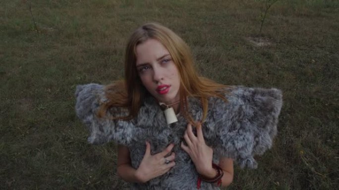 近距离拍摄金发女孩的肖像，她穿着一种异教动物般的毛皮，由皮肤制成，站在草地背景下，抬头仰望天空，风吹