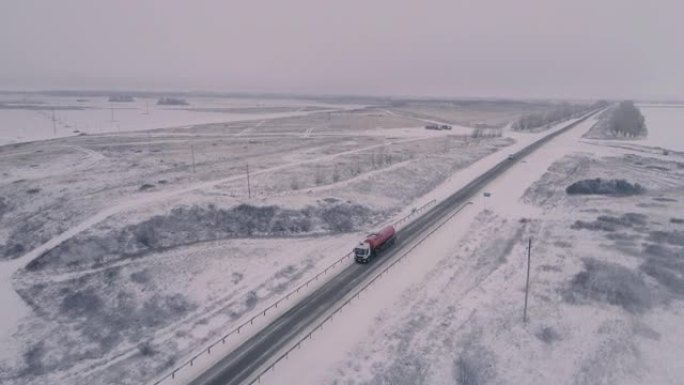 积雪覆盖的冬季高速公路的俯视图。卡车司机开着一辆红色油轮。高高的光秃秃的树木。雪场上的电线。朦胧的天