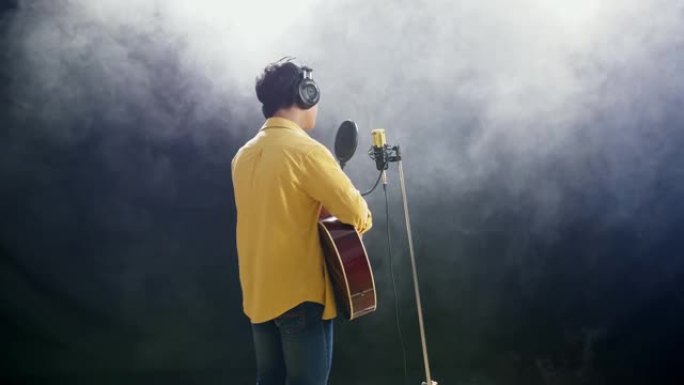 一个戴着耳机的小男孩的后视图弹吉他，在白色烟雾黑色背景上对着电容式麦克风唱歌