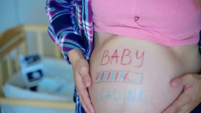 孕妇在准备好的婴儿床背景下抚摸着她怀孕的腹部，粉红色和蓝色的婴儿装载铭文画在女人的肚子上，等待婴儿到