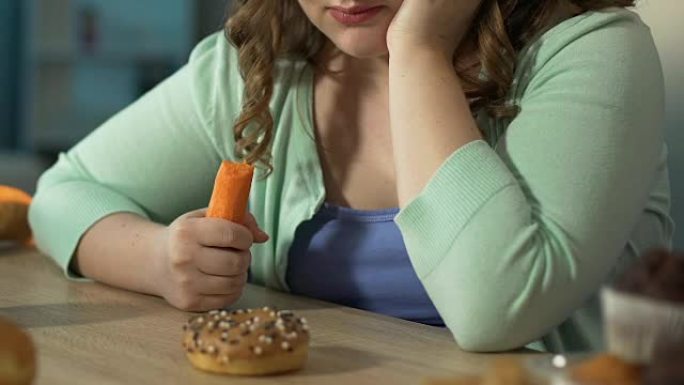 超重的女孩在吃新鲜胡萝卜时悲伤地看着桌子上的甜甜圈