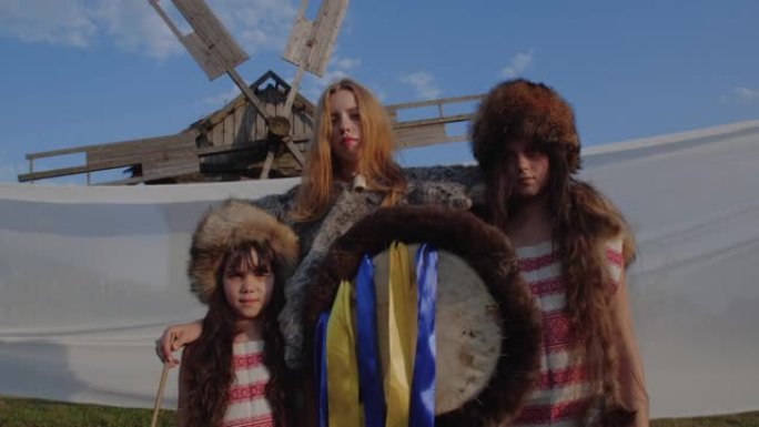 时尚的肖像，年轻女子留着红色长发，两个女孩穿着皮草套装，手里拿着萨满铃鼓站着，在旧木制风车的背景下看