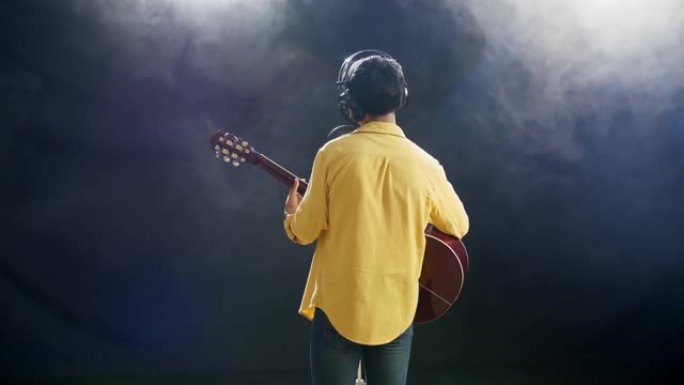 一个戴着耳机的小男孩的后视图弹吉他，在白色烟雾黑色背景上对着电容式麦克风唱歌