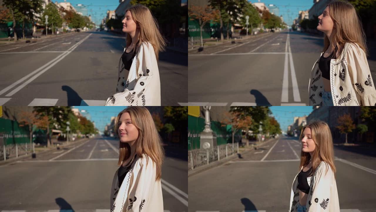 自信美丽的少女穿着刺绣衬衫微笑慢动作穿越城市街道的侧视图。快乐少年在阳光下右边行走的跟踪镜头。