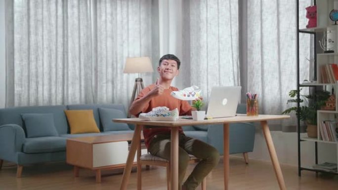 微笑的亚洲男孩鞋类设计师拿着彩色图案运动鞋，在家里的笔记本电脑上工作时向相机竖起大拇指