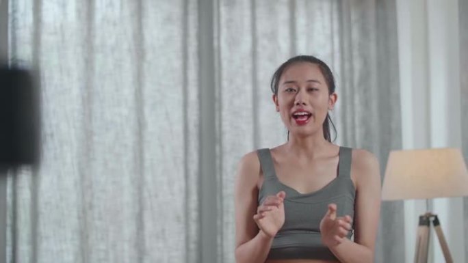 年轻的亚洲教练女性穿着运动服对着镜头说话，一边在家录制教学练习