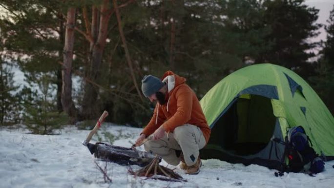一名男子用刀准备木棉，以便在日落时在冬季森林中生火。冬季生存概念。