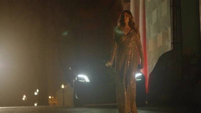 晚上，穿着金色连衣裙的美丽奢华女人在车灯前慢慢行走