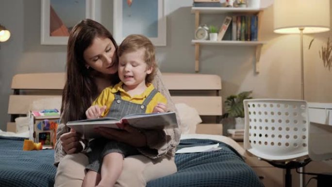 妈妈和宝宝阅读。妈妈和2-3岁的孩子晚上坐在床上的卧室里看书，女人和男孩用手指指着书中的东西。慢动作