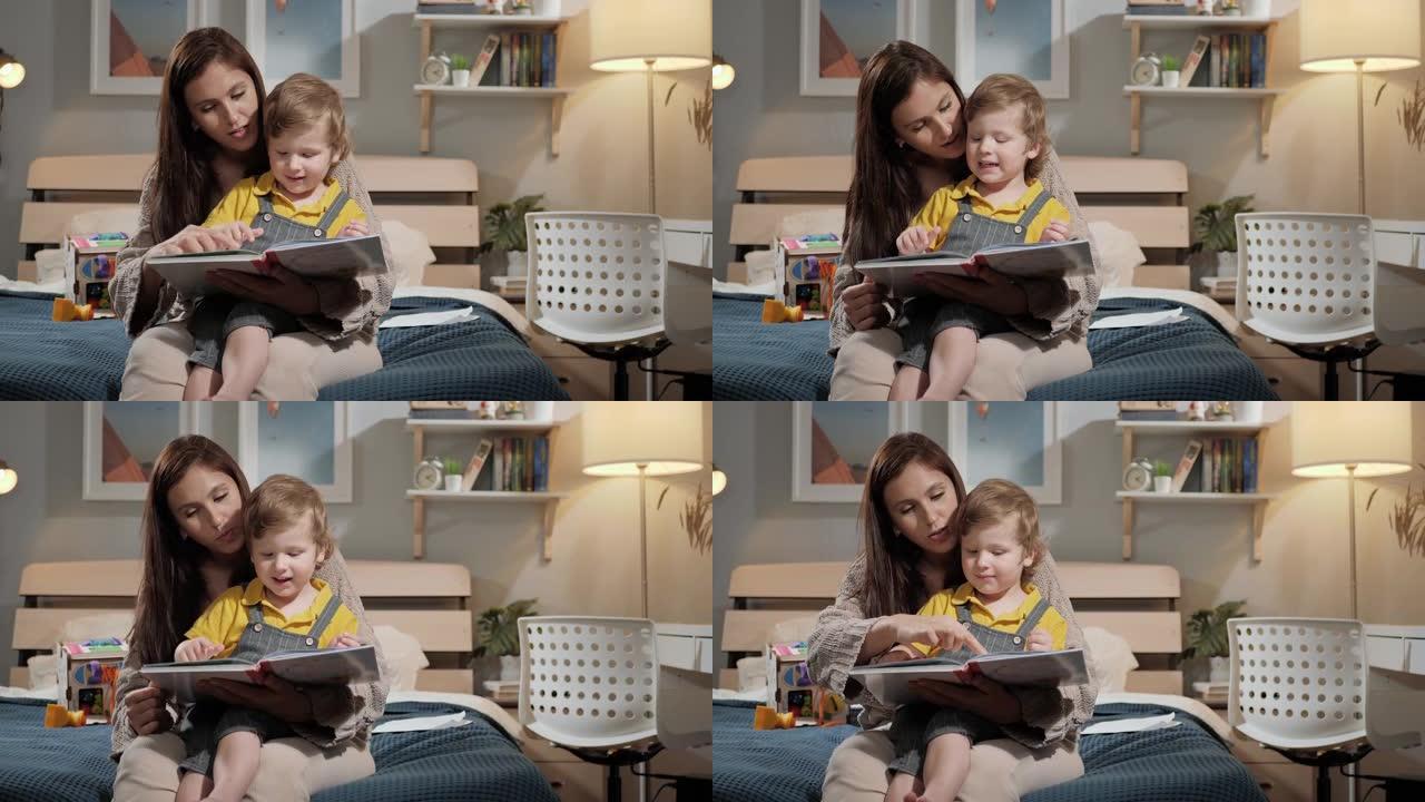 妈妈和宝宝阅读。妈妈和2-3岁的孩子晚上坐在床上的卧室里看书，女人和男孩用手指指着书中的东西。慢动作