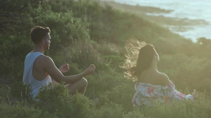 两个朋友坐在海边的草崖上放松身心。莲花姿势冥想并练习健康