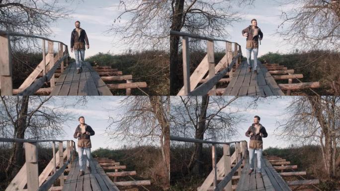 年轻人沿着古老的木吊桥走小。背包徒步旅行的旅行者在秋天的风景