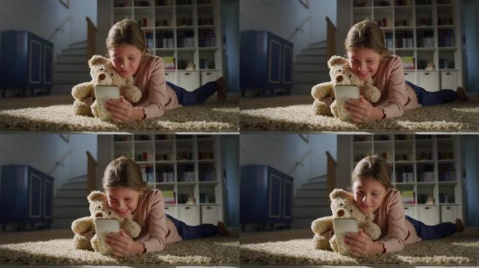 一个小女孩的肖像抱着她的泰迪熊，并在客厅的智能手机上观看视频。快乐可爱的女孩使用手机，玩手机教育游戏