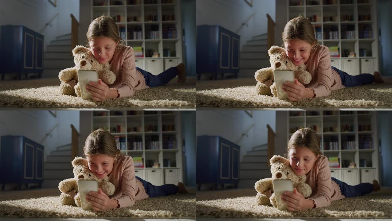 一个小女孩的肖像抱着她的泰迪熊，并在客厅的智能手机上观看视频。快乐可爱的女孩使用手机，玩手机教育游戏
