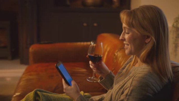 在家中的女人坐在沙发上带火用蓝屏手机拿着一杯酒