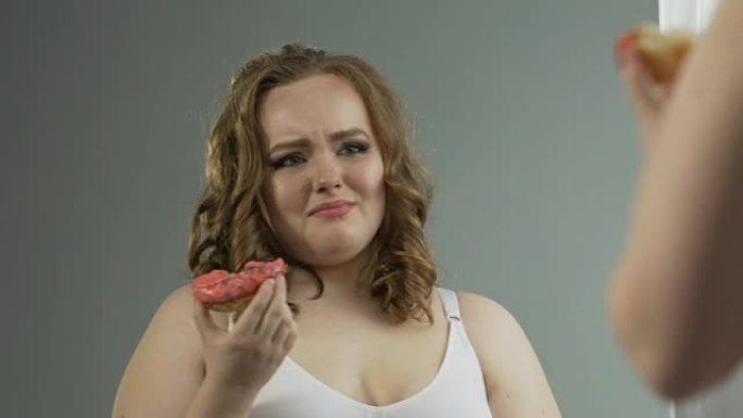 沮丧的超重女孩看着镜子里的身体，绝望地吃甜甜圈