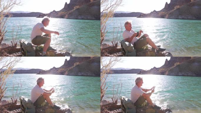 男性徒步旅行者在湖岸放松，使用电子阅读器