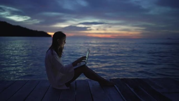 日落时在甲板上的笔记本电脑上工作的年轻女子