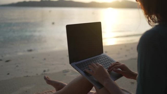 日落时，一名年轻女子在海滩上的笔记本电脑上工作的特写镜头