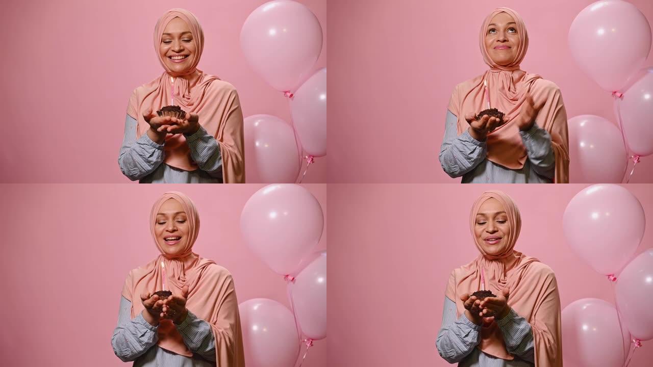 美丽的穆斯林妇女在粉红色的头巾，许下美好的愿望，吹灭蜡烛在生日蛋糕