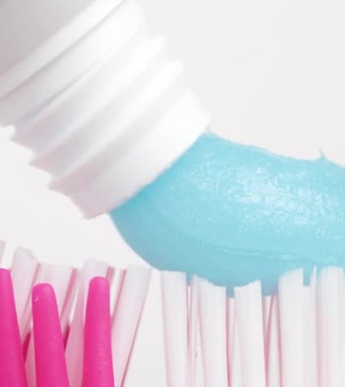牙刷和牙膏。白色背景上挤压蓝色牙膏的粉红色牙刷的垂直宏观视图。慢动作