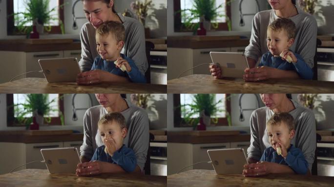 母亲和儿子在家里的厨房里用数字平板电脑拍摄的真实照片。男性蹒跚学步的孩子在享受零食的同时学习如何使用