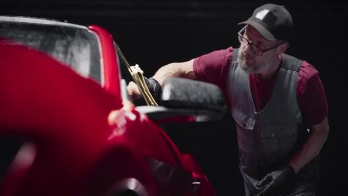 成人汽车美容师穿着制服洗红色跑车，用超细纤维毛巾清洁车辆。在时尚的美国汽车上工作的技术人员。广告的商