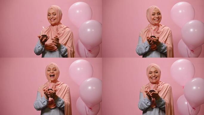 穆斯林妇女在粉红色的头巾，微笑着，唱着生日快乐的歌，拿着纸杯蛋糕和点燃的蜡烛