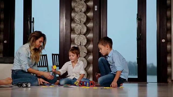 快乐的母亲和两个孩子坐在他的乡间别墅的地板上，花时间在一起收集儿童设计师。带全景窗户的客厅