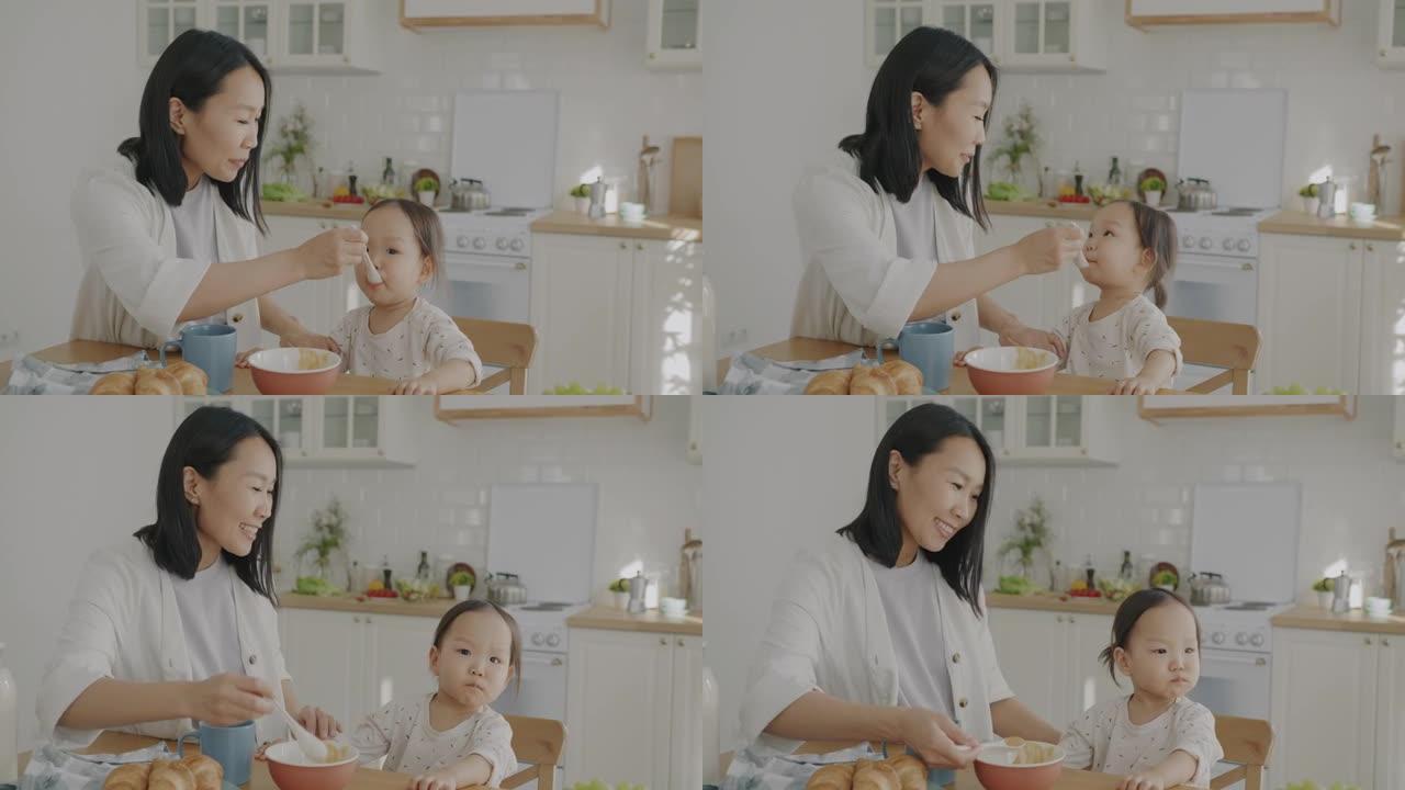 快乐的亚洲女士用婴儿食物喂养小女儿，微笑着看着公寓厨房里的孩子
