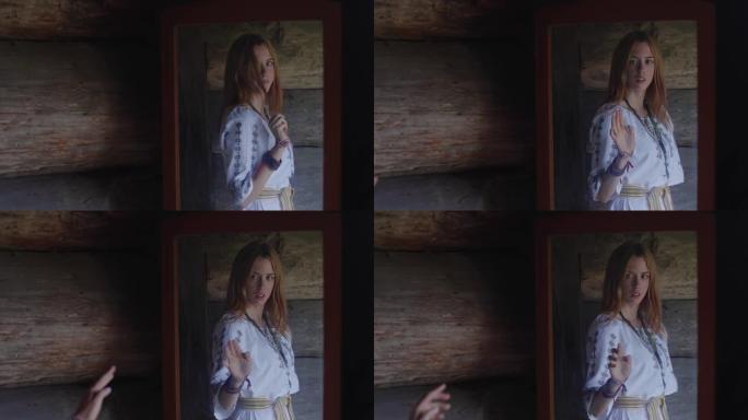 穿着刺绣衬衫的迷人年轻金发女子看着一间旧木屋里的镜子，微笑着伸出手触摸她的倒影。占卜、神秘主义、魔法