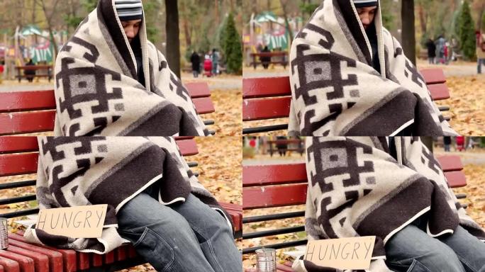 年轻的无家可归者坐在公园的长椅上，寻求帮助，饥饿，贫困