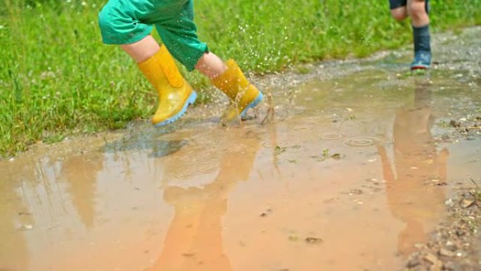 SLO MO两个穿着雨靴的男孩在阳光下穿过泥泞的水坑