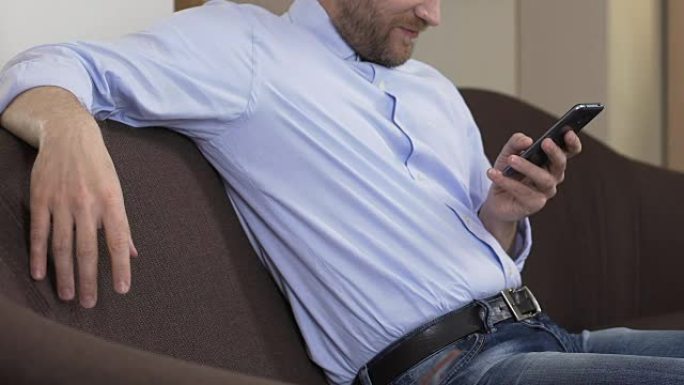 大胡子男人坐在沙发上滚动智能手机，社交网络，小工具