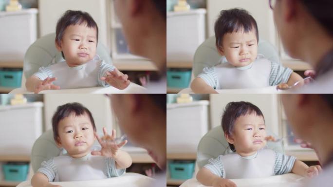 年轻的亚洲男婴在家里的午餐时间坐在椅子上时，母亲哭着拒绝吃饭或食物。儿童日常生活方式照顾或幼儿营养食