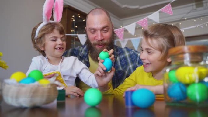 复活节家庭传统。父亲和两个白种人快乐的孩子，兔子耳朵在家里坐在一起的桌子上玩复活节彩蛋，用油漆过节。