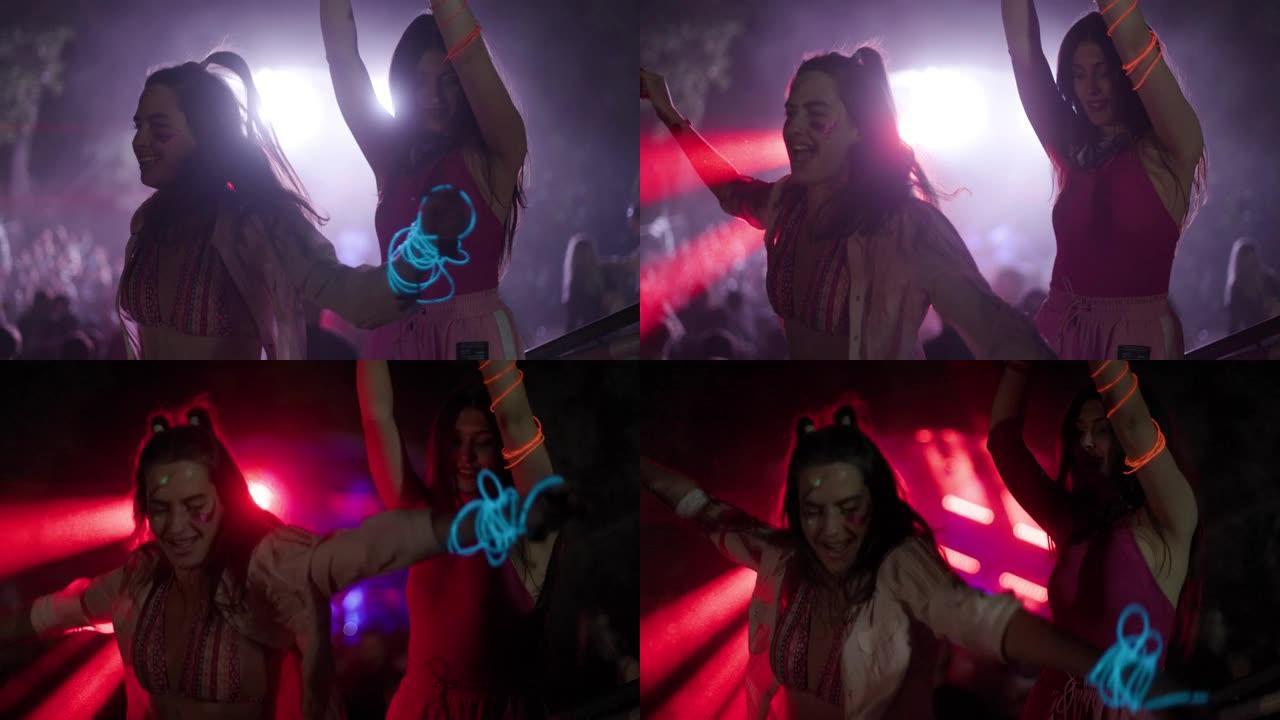 两个快乐的年轻女孩在闪烁的灯光和激光的开放空间派对上跳舞。背景中的焦点人群和dj舞台。