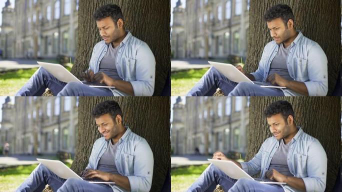 跨国年轻人坐在树下，在笔记本电脑上打字，微笑，聊天