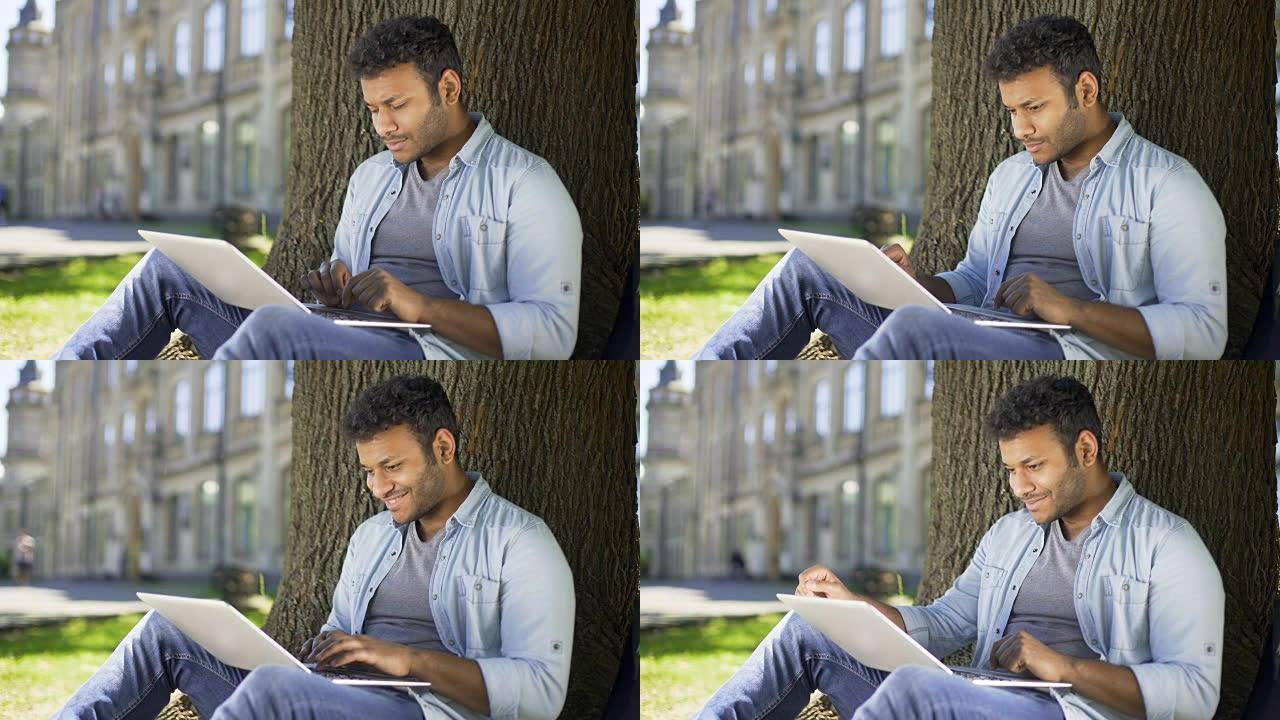 跨国年轻人坐在树下，在笔记本电脑上打字，微笑，聊天