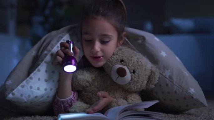 睡前给她最喜欢的泰迪熊看书的小女孩的肖像。快乐可爱的女孩玩她的玩具，用手电筒，讲故事。童年和想象力的