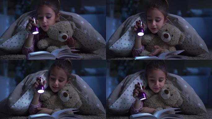 睡前给她最喜欢的泰迪熊看书的小女孩的肖像。快乐可爱的女孩玩她的玩具，用手电筒，讲故事。童年和想象力的