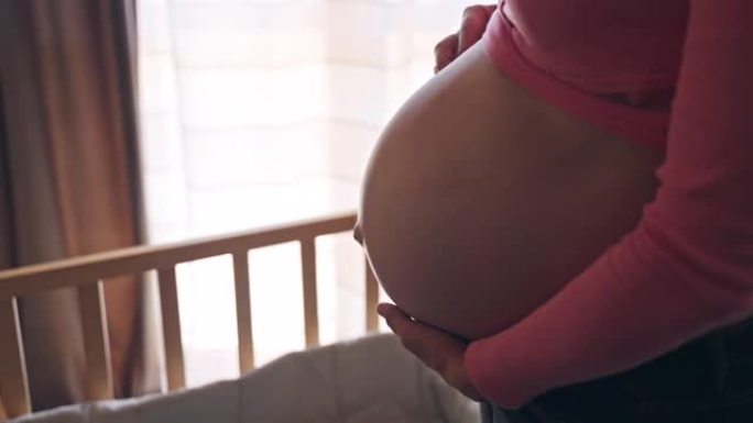 美丽的孕妇用手抚摸她的肚子站在木制婴儿床附近的房间里，未来的妈妈在肚子里展示她对婴儿的真正爱和感情，