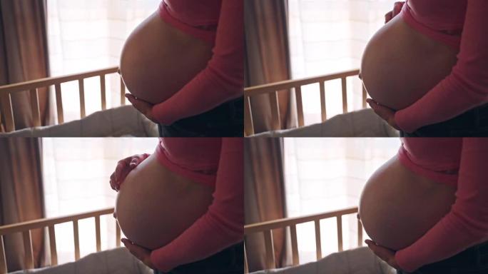 美丽的孕妇用手抚摸她的肚子站在木制婴儿床附近的房间里，未来的妈妈在肚子里展示她对婴儿的真正爱和感情，