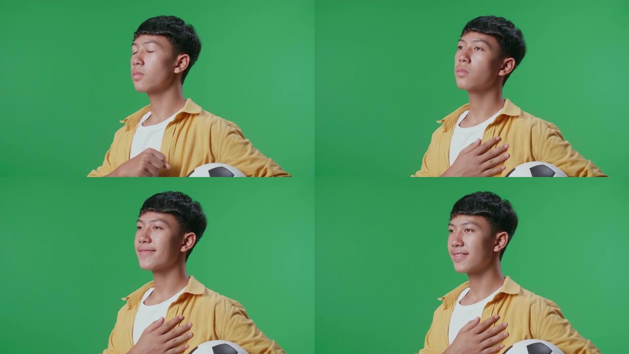 亚洲男孩的近距离侧视图，将球放在他的心脏上，以表示敬意，同时在绿屏背景上欢呼足球