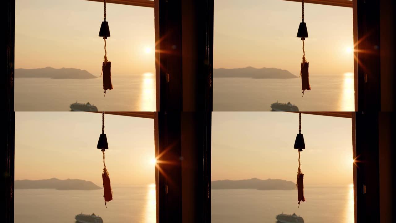 风铃挂在窗户上，在背景中，海洋或海洋上有美丽的日落，阳光照进相机。门口挂着带音符的铃铛。