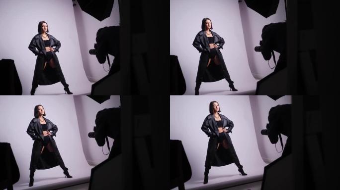 性感年轻的时尚女模特在黑色泳装和皮靴和外套的工作室与摄影师在白色背景下拍摄。后台。