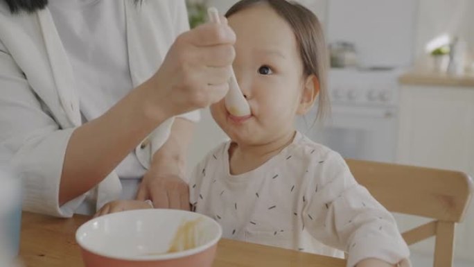 可爱的孩子一边吃婴儿食品，一边照顾母亲在家里的厨房里用果泥喂养女儿